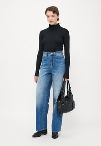 Woman wearing black turtleneck, high-waisted blue jeans, black shoes, holding black handbag, standing against plain light background.