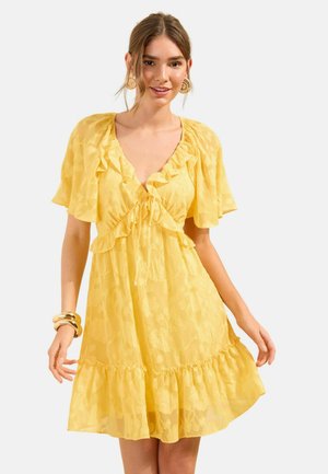 Kobieta ubrana w krótką żółtą sukienkę w kwiaty z falbankowymi rękawami i detalami, uzupełniona dużymi złotymi kolczykami i bransoletkami.