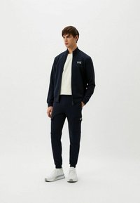 Giacca zip-up navy con colletto alto, pantaloni cargo navy abbinati, entrambi realizzati in stoffa morbida. Sneakers bianche con dettagli grigi. Logo del marchio visibile.