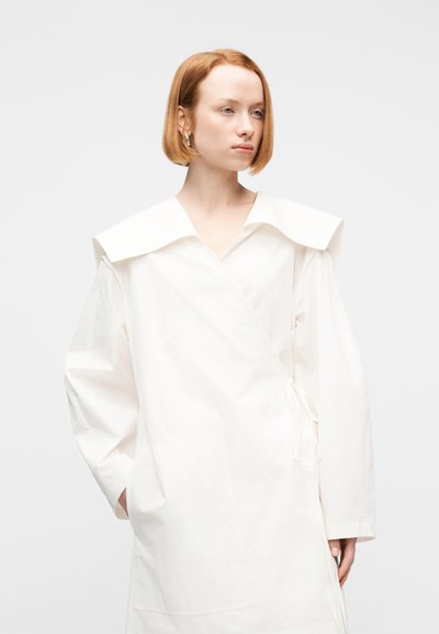 Γυναίκα με κοντά κόκκινα μαλλιά, φορώντας ένα άνετο λευκό φόρεμα τύπου wrap με φαρδιά γιακά, μπροστά σε απλό λευκό φόντο.