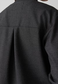 Gros plan de l'arrière d'une personne portant une veste en tissu texturé gris foncé avec un pli central dans le dos et des coutures visibles.