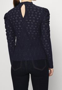 Námořnická modrá halenka s dlouhým rukávem a texturovanými diamantovými vzory, s nařasenými rameny a detailem ve tvaru klíčové dírky na zadní straně. Vyrobena z prodyšného materiálu.