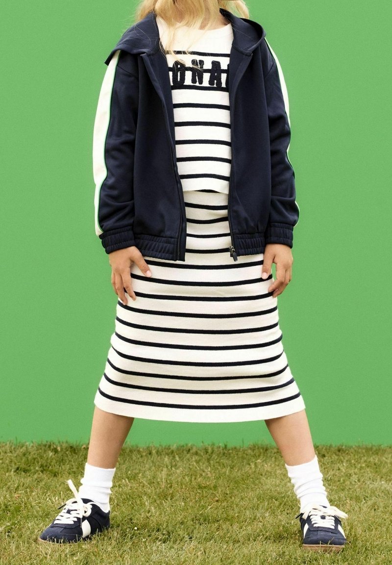 Enfant portant une robe rayée noire et blanche avec un texte, une veste bleu marine à rayures blanches, des chaussettes blanches et des baskets bleu marine, debout sur l'herbe.