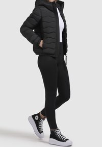 Czarna kurtka puchowa z kapturem, pikowany wzór, zamek błyskawiczny z przodu, w połączeniu z czarnymi legginsami i czarnymi sneakersami za kostkę.