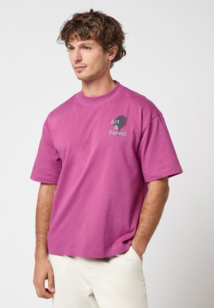 Jonge man met krullend haar, draagt een losse magenta T-shirt met de tekst "Art & Forest" en witte broek, handen in de zakken, kijkt zijwaarts.