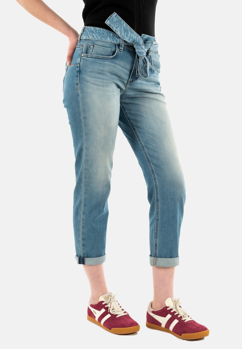 Jeans in denim blu chiaro a cropped con una cintura annodata a fantasia, dotati di tasche e risvolti, abbinati a sneakers bordeaux e bianche.