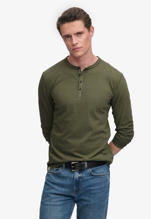 Jeune homme aux cheveux courts bruns, portant un t-shirt henley à manches longues couleur olive et un jean bleu, debout les mains derrière le dos.