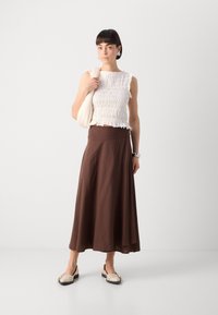 Top corto bianco con smock e orlo a frangia, gonna marrone lunga fino alla caviglia e scarpe slip-on color crema con dettagli decorativi. Borsa a tracolla neutra.
