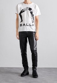 Vit t-shirt med en svart grafisk design och ordet "BALLY" längst ner. Kombineras med åtsittande svarta läderbyxor och svarta sneakers.