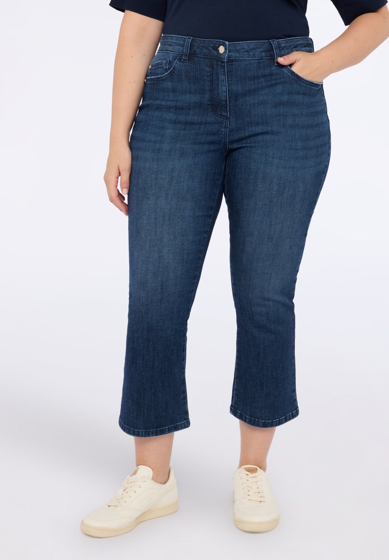 Tummansininen farkkuhousut, joissa on korkea vyötärö ja lyhennetty helma, edessä taskut ja nappikiinnitys, yhdistettynä beigeihin lenkkitossuihin.