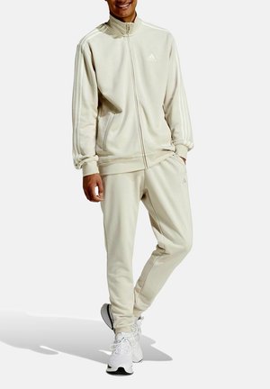 Tuta beige realizzato in tessuto morbido; giacca con zip e tre strisce bianche, abbinato a pantaloni. Caratterizzato da una vestibilità comoda e polsini elasticizzati.