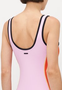 Donna che indossa un costume da bagno smanicato con un design a blocchi di colore, rosa, arancione e nero, vista da dietro su uno sfondo neutro.