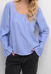 Blouse rayée bleu clair en tissu doux, avec un col rond et de longues manches amples avec un détail plissé, associée à un jean noir.