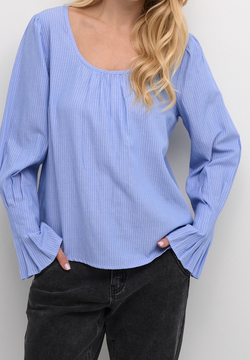 Blouse rayée bleu clair en tissu doux, avec un col rond et de longues manches amples avec un détail plissé, associée à un jean noir.