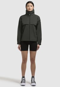 Giacca zip-up verde scuro con colletto alto, tasca frontale e due tasche laterali; indossata sopra pantaloni corti neri e scarpe alte.