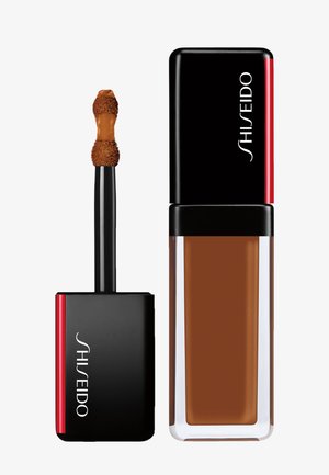 Shiseido liquid foundation in a rectangular clear bottle with black and red cap, applicator wand with brown liquid foundation on tip.