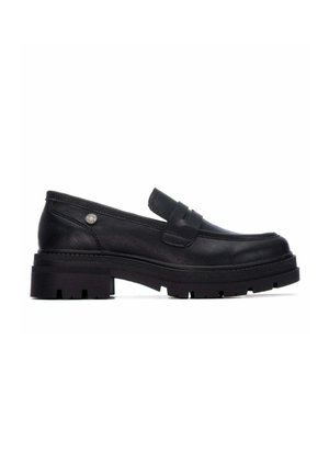 Mocasín negro de cuero con suela gruesa de tacos y tacón bajo, con una tira en la parte superior y un pequeño emblema metálico redondo cerca del tacón.