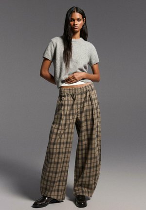 Donna in piedi davanti a uno sfondo grigio, indossa un maglione grigio a maniche corte, pantaloni larghi a quadri marroni e scarpe nere.