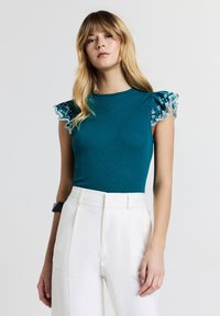Top teal a coste con maniche in pizzo trasparente a volant; abbinato a pantaloni bianchi a vita alta con una finitura liscia e testurizzata.