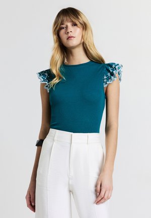 Top teal a coste con maniche in pizzo trasparente a volant; abbinato a pantaloni bianchi a vita alta con una finitura liscia e testurizzata.