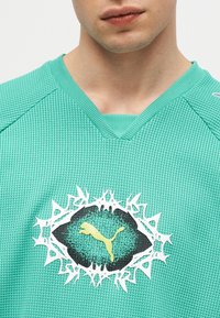 Κοντινό πλάνο ενός ατόμου που φοράει μπλουζάκι σε μέντα πράσινο με υφή, με κίτρινο λογότυπο Puma μέσα σε μαύρο και άσπρο κυκλικό σχέδιο με ακίδες.