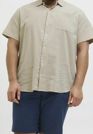 Chemise beige à manches courtes avec boutonnière et poche poitrine, associée à un short bleu marine longueur genoux porté par une personne debout.