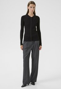 Mujer con un suéter negro de botones ajustado y de canalé, pantalones de pierna ancha grises con pliegues y zapatos negros de punta, de pie frente a un fondo neutro.