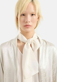 Foulard di seta legato al collo, color crema, con una texture liscia. Indossato con una giacca bomber di colore chiaro caratterizzata da un design con un motivo sottile.