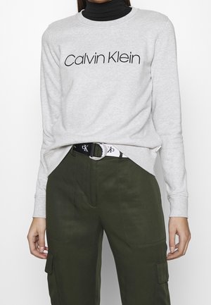 Personne portant un sweat-shirt gris clair Calvin Klein par-dessus un col roulé noir, un pantalon cargo vert olive et une ceinture Calvin Klein noire et blanche.