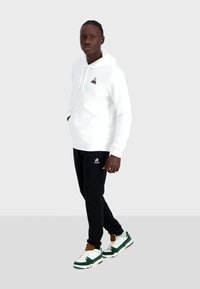 Felpa bianca in cotone con tasca frontale e logo ricamato, abbinata a pantaloni jogger neri con logo coordinato, completata da sneaker verdi e bianche.