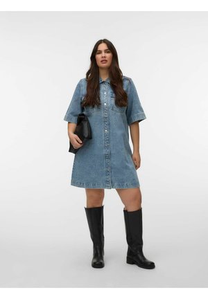 Kobieta w sięgającej do kolan, zapinanej na guziki sukience z denimu, czarnych wysokich botkach do kolan, trzymająca czarną kopertówkę, stojąca na tle jednolitej białej ściany.