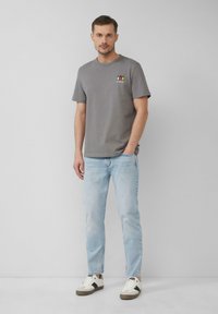 Szara bawełniana koszulka z okrągłym dekoltem, z małym, wielokolorowym nadrukiem na piersi, w zestawieniu z jasnobeżowymi dżinsami i trampkami.