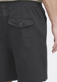 Shorts noirs avec une taille élastique et une poche arrière avec bouton. Le matériau semble doux avec une texture lisse et un design simple.