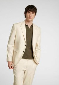 Kremowy garnitur z dwoma czarnymi guzikami, noszony na ciemnozielonej koszulce z dekoltem w serek. Gładka tekstura tkaniny, dopasowany krój i wyrafinowany design.