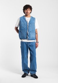 Chaleco de mezclilla con cuatro bolsillos, cremallera frontal, de color azul claro. Lleva una camiseta blanca debajo, combinada con pantalones de mezclilla azul a juego y zapatos negros.