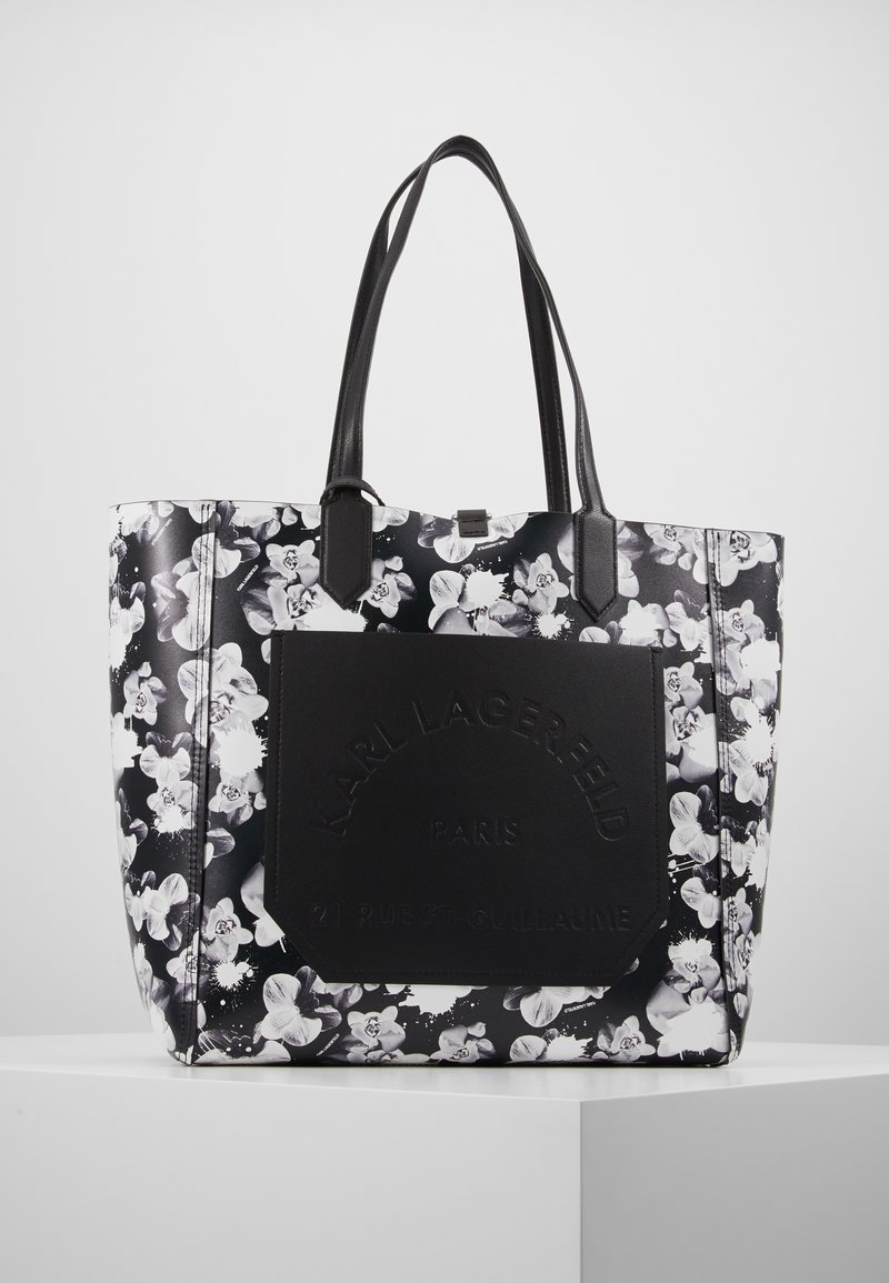 Sac cabas floral noir et blanc avec des accents en cuir noir, présentant un panneau logo texturé et deux bretelles d'épaule.