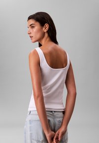 Débardeur blanc côtelé à larges bretelles et dos carré, porté avec un jean en denim bleu clair taille haute, mains jointes dans le dos.