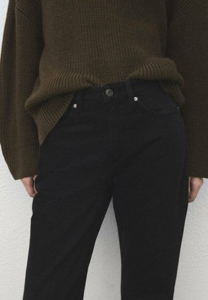 Približna snimka osobe koja nosi maslinasto zeleni debeli džemper uvučen u crne hlače visokog struka s džepovima i gumbima.