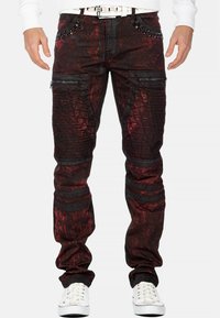 Pantaloni da uomo rossi e neri con effetto usurato, tasche con zip, dettagli testurizzati e borchie sulla vita. Materiale in misto cotone.