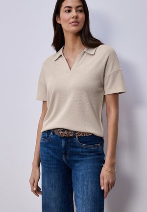 Kobieta ubrana w beżowy top z krótkim rękawem i kołnierzykiem, wpuszczony w niebieskie dżinsy z paskiem w panterkę, stojąca na tle jasnoszarej ściany.