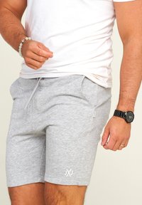 Graue Baumwollshorts mit einem elastischen Bund, Seitentaschen und einem weißen Logo. Kombiniert mit einem kurzärmeligen weißen Hemd und einer schwarzen Uhr.
