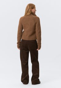 Personne portant un pull col roulé marron et un pantalon en velours côtelé marron foncé, se tenant de dos sur un fond clair.