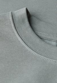 Detailní záběr na složenou šedou mikinu s kulatým výstřihem ukazující žebrovaný límec a jemnou texturu pleteniny.