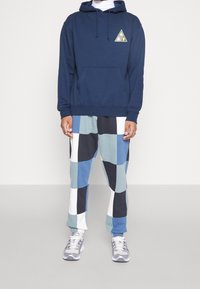 Sudadera con capucha azul marino con un logo de triángulo verde, combinada con pantalones de patchwork multicolores en azul, negro y blanco. Zapatillas grises completan el conjunto.