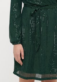Main reposant à côté de la taille portant une robe vert foncé à sequins avec une ceinture en tissu nouée et des manches longues transparentes.