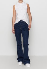 Bílá bezrukávová polo košile s límečkem a knoflíkovým zapínáním, v kombinaci s tmavě modrými zvonovými džíny a bílými teniskami. Jednoduchý a pohodlný design.