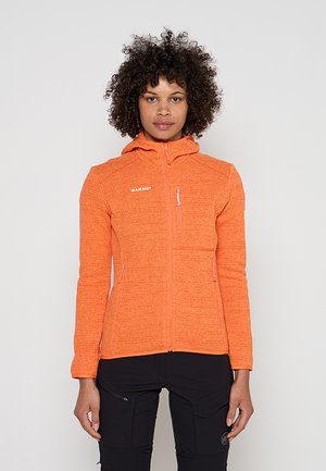 Donna con capelli ricci che indossa una felpa con zip arancione Mammut e pantaloni neri, in piedi davanti a uno sfondo semplice.