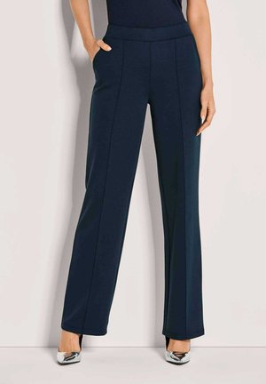 Vrouw in marineblauwe wijde broek met voornaad, met één hand in de zak, gecombineerd met zilveren puntige hakken tegen een effen achtergrond.