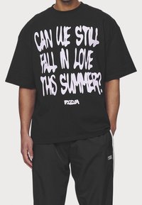 Svart oversized t-shirt med fet vit text som står: "KAN VI FORTFARANDE FALLA I KÄRLEK DETTA SOMMAR?" i en droppande fontdesign.