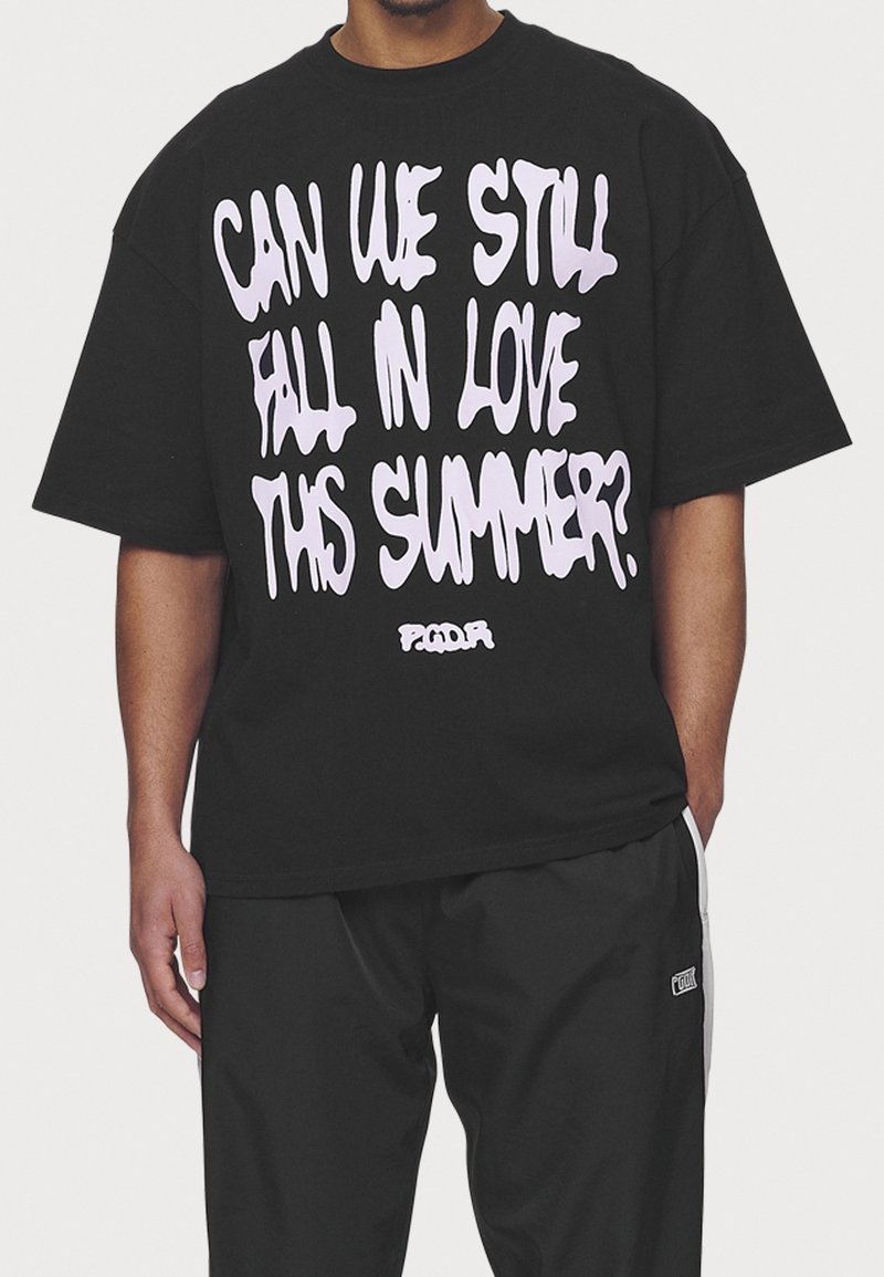 Svart oversized t-shirt med fet vit text som står: "KAN VI FORTFARANDE FALLA I KÄRLEK DETTA SOMMAR?" i en droppande fontdesign.